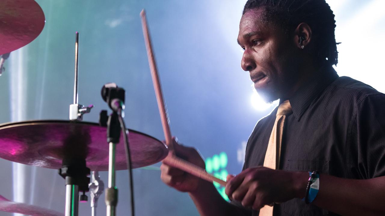Ein Mann mit schwarzen Haaren sitzt auf einer Bühne hinter einem Schlagzeug und spielt.