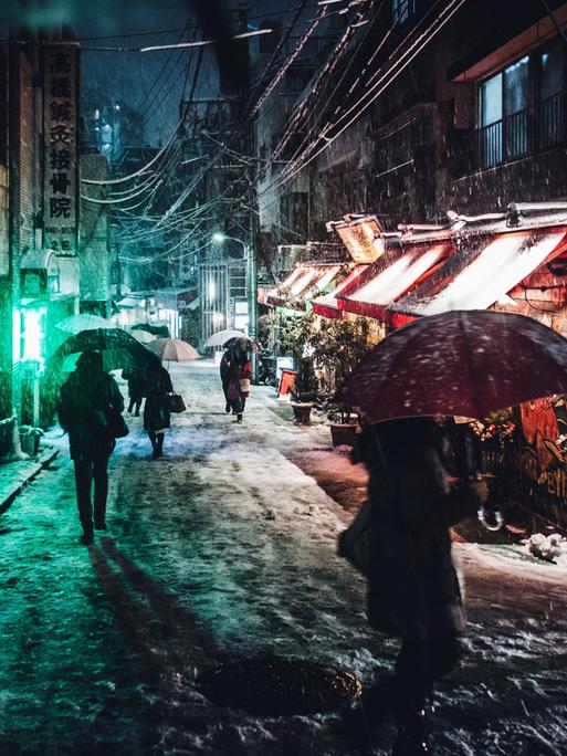 Menschen mit Regenschirmen gehen abends im Schirm durch eine kleine Straße in Tokio.