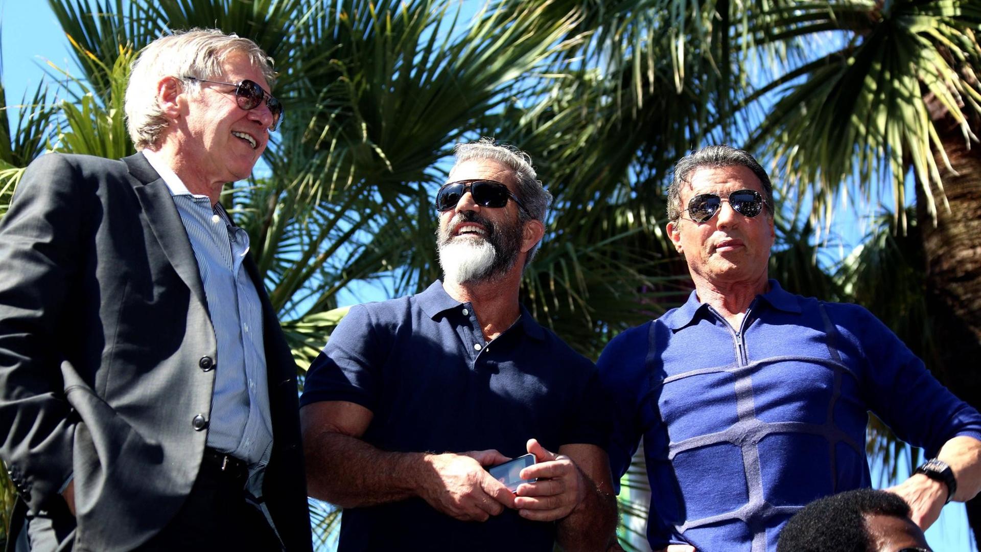 Harrison Ford, Mel Gibson und Sylvester Stallone stehen in Cannes vor Palmen.