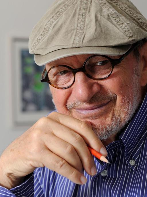 Das Bild zeigt den Cartoonist Guillermo Mordillo mit freundlichem Blick in Richtung Kamera. Er trägt eine Brille, einen Hut und ein blau-weiß kariertes Hemd. In seiner rechten Hand hält er einen Bleistift und stützt sich am Kinn ab.