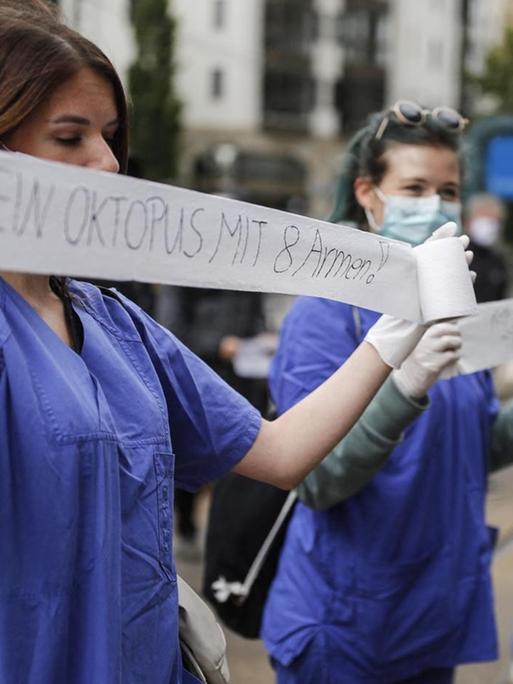 Eine medizinische Pflegekraft, in blauer Krankenhaus-Bekleding, hält während einer Demonstration ein Stück Toilettenpapier mit der Aufschrift "Ich bin kein Oktopus mit 8 Armen" in den Händen.