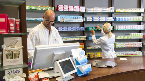 Ein Apotheker steht hinter der Ladentheke und schaut in den Computer, hinter nimmt eine Pharmazeutische Assistentin ein Medikament aus dem Regal