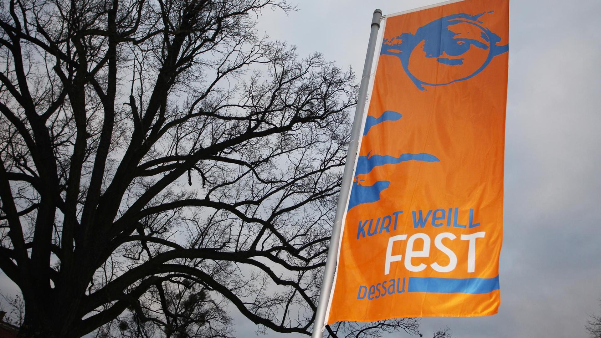Eine orangefarbene Fane mit hellblauer Schrift weht im Wind vor einem großen, kahlen Baum.
