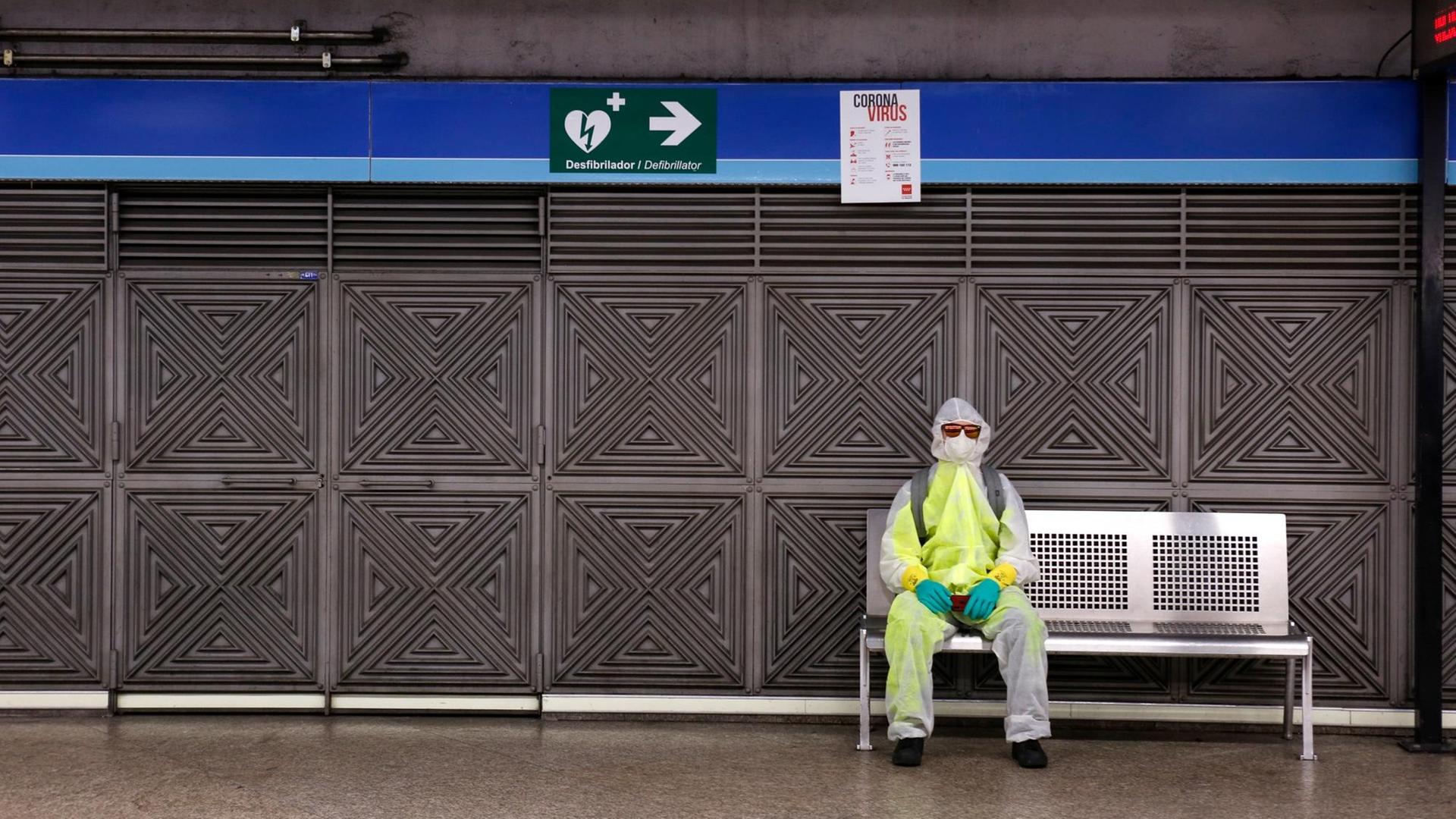 Ein Mann mit Schutzkleidung und Mundschutz sitzt in einer Metrostation in Madrid auf einer Bank.