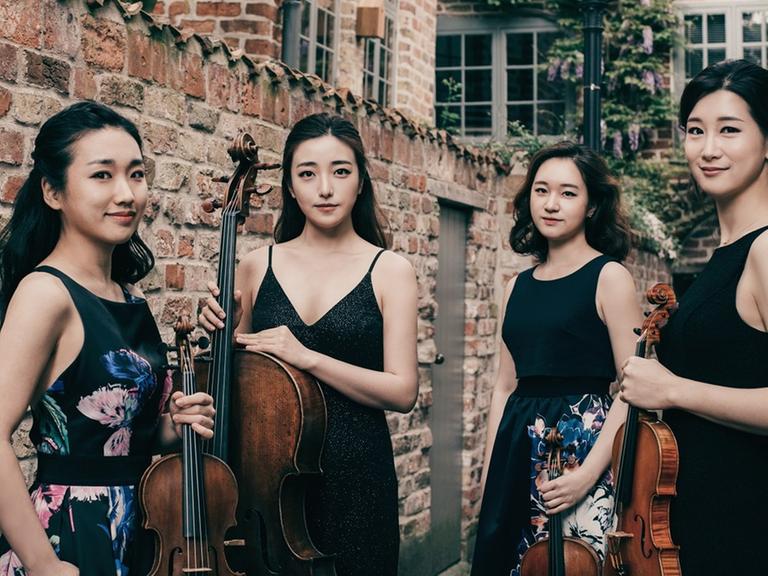 Die vier Musikerinnen stehen in blumenbedruckten schwarzen Konzertkleidern vor einer Ziegelmauer