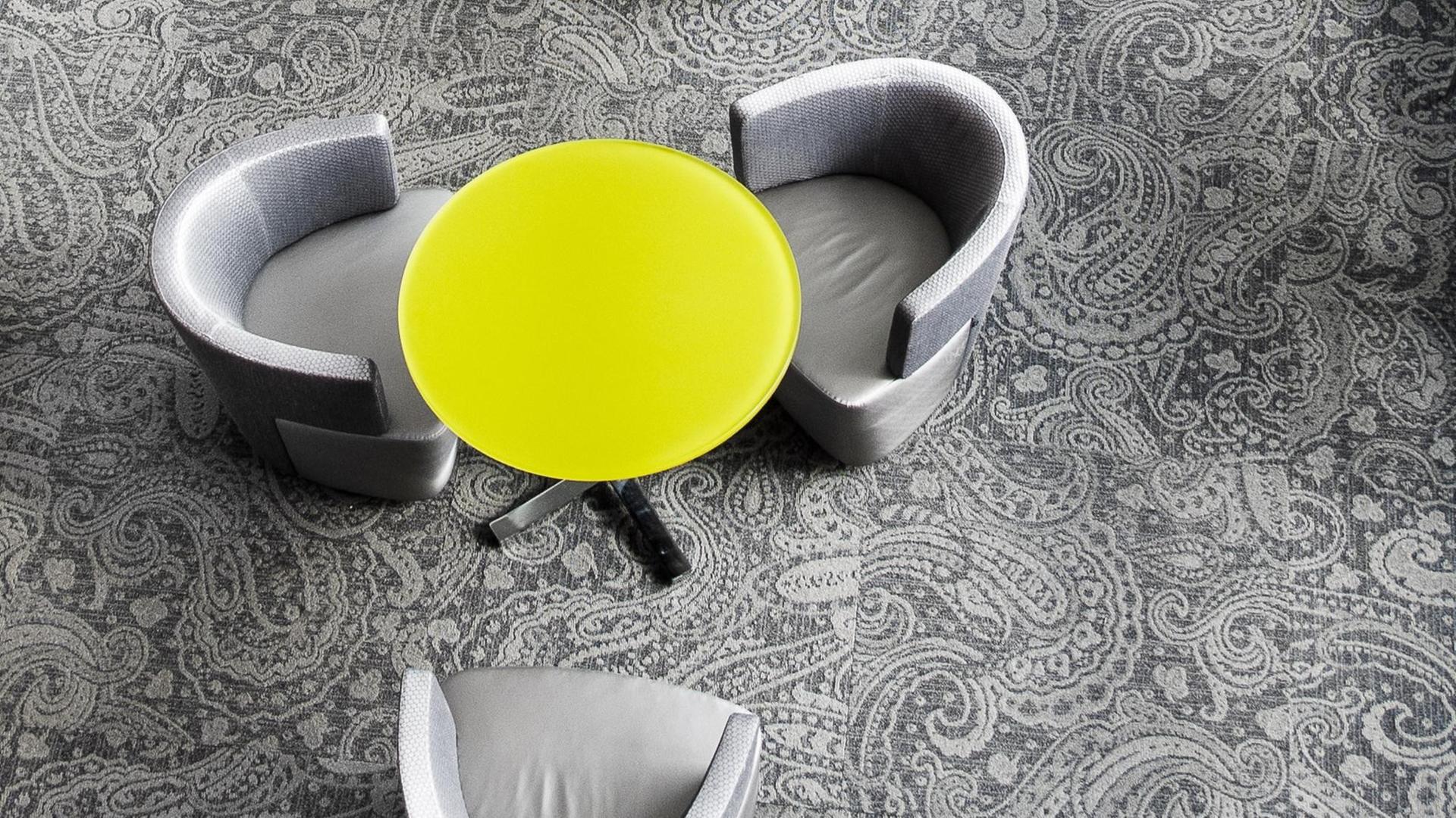Ein gelber runder Tisch auf grauem Teppich inmitten von grauen Stühlen.