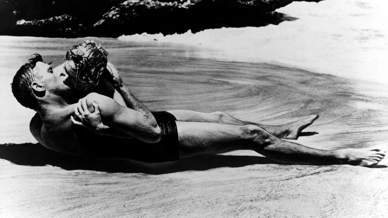 
Ein Film-Still zeigt die Schauspielerin Deborah Kerr und Filmpartner Burt Lancaster bei einem Kuss am Strand von Hawaii von einer Welle umspült