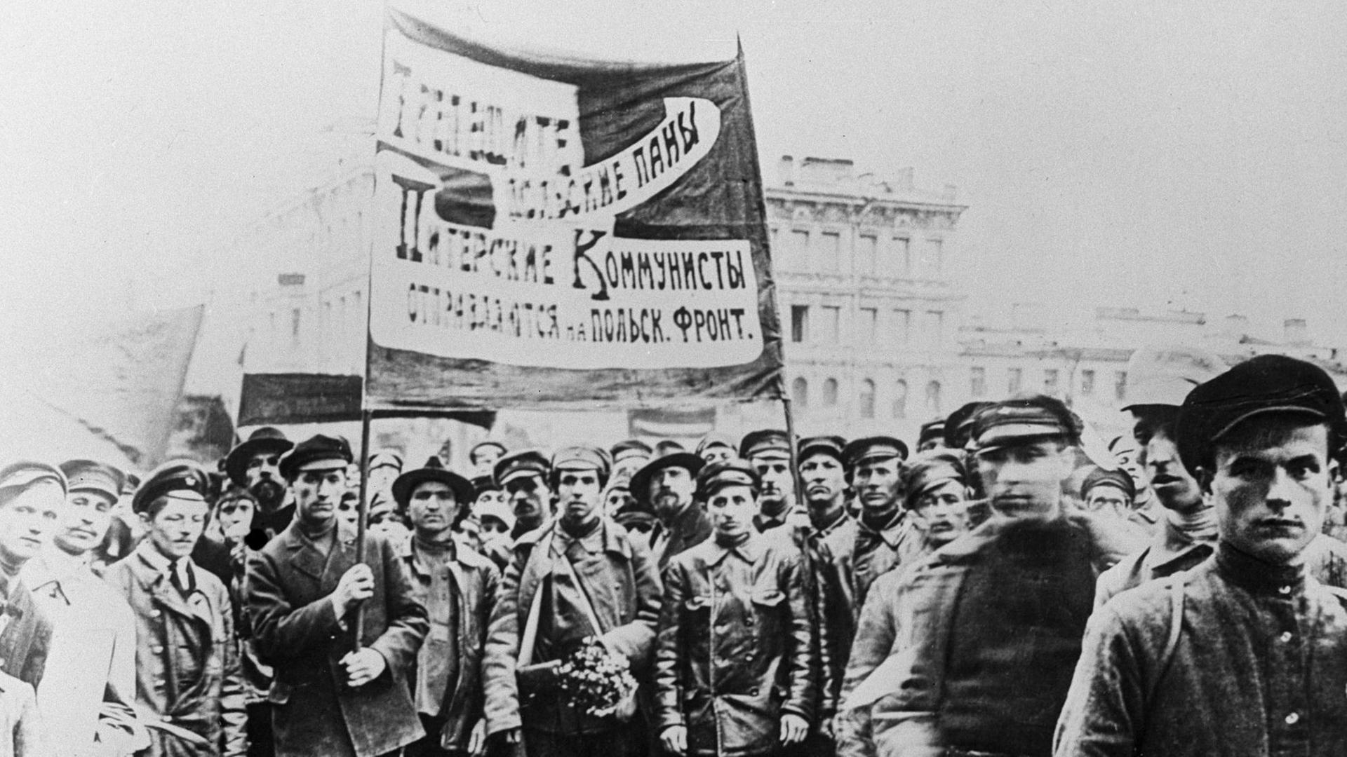 Ein Schwarzweiß-Foto zeigt etwa ein Dutzend Männer, teils in Uniform zumeist in Lederjacken und mit Schirmmützen ein großes, kyrillisch beschriebenes Banner hochhaltend
