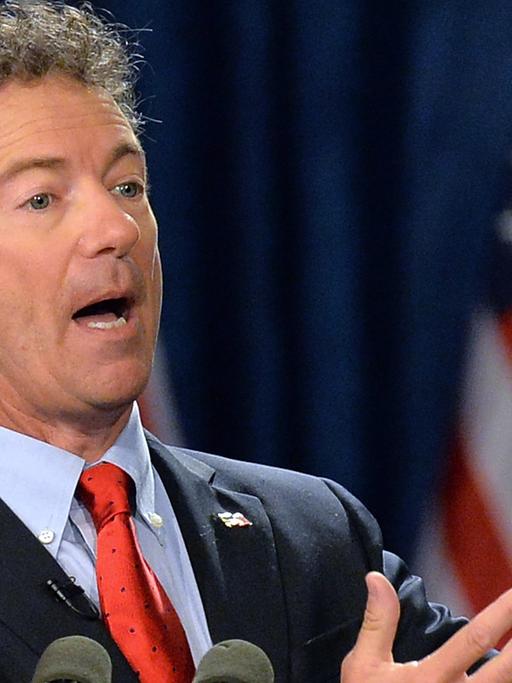 Rand Paul an einem Rednerpult sprechend und gestikulierend, im Hintergrund amerikanische Flaggen.