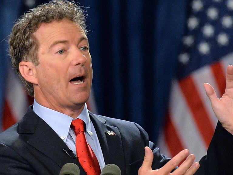 Rand Paul an einem Rednerpult sprechend und gestikulierend, im Hintergrund amerikanische Flaggen.