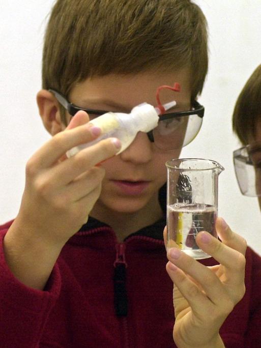 Ein achtjähriger Junge träufelt Kokosfett in ein Gefäß mit Leitungswasser, zwei andere Jungs schauen ihm dabei zu. Die Kinder tragen Schutzbrillen.