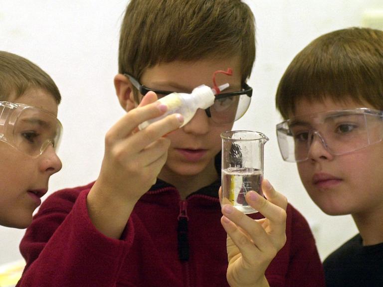 Ein achtjähriger Junge träufelt Kokosfett in ein Gefäß mit Leitungswasser, zwei andere Jungs schauen ihm dabei zu. Die Kinder tragen Schutzbrillen.