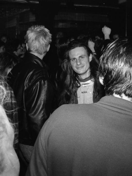 Schwarzweißfoto von der Tanzfläche in einer Bar oder Discothek. Menschen unterhalten sich, mittendrin steht ein Mann und schaut in Richtung der Fotografin. Es ist der Schriftsteller Rainald Goetz.