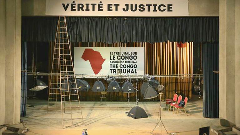 Die Ansicht einer Bühne, über der ein Plakat "Verité et Justice" schwebt, hinten eine Karte des Kongo mit Schrift "Das Kongo Tribunal" in drei Sprachen