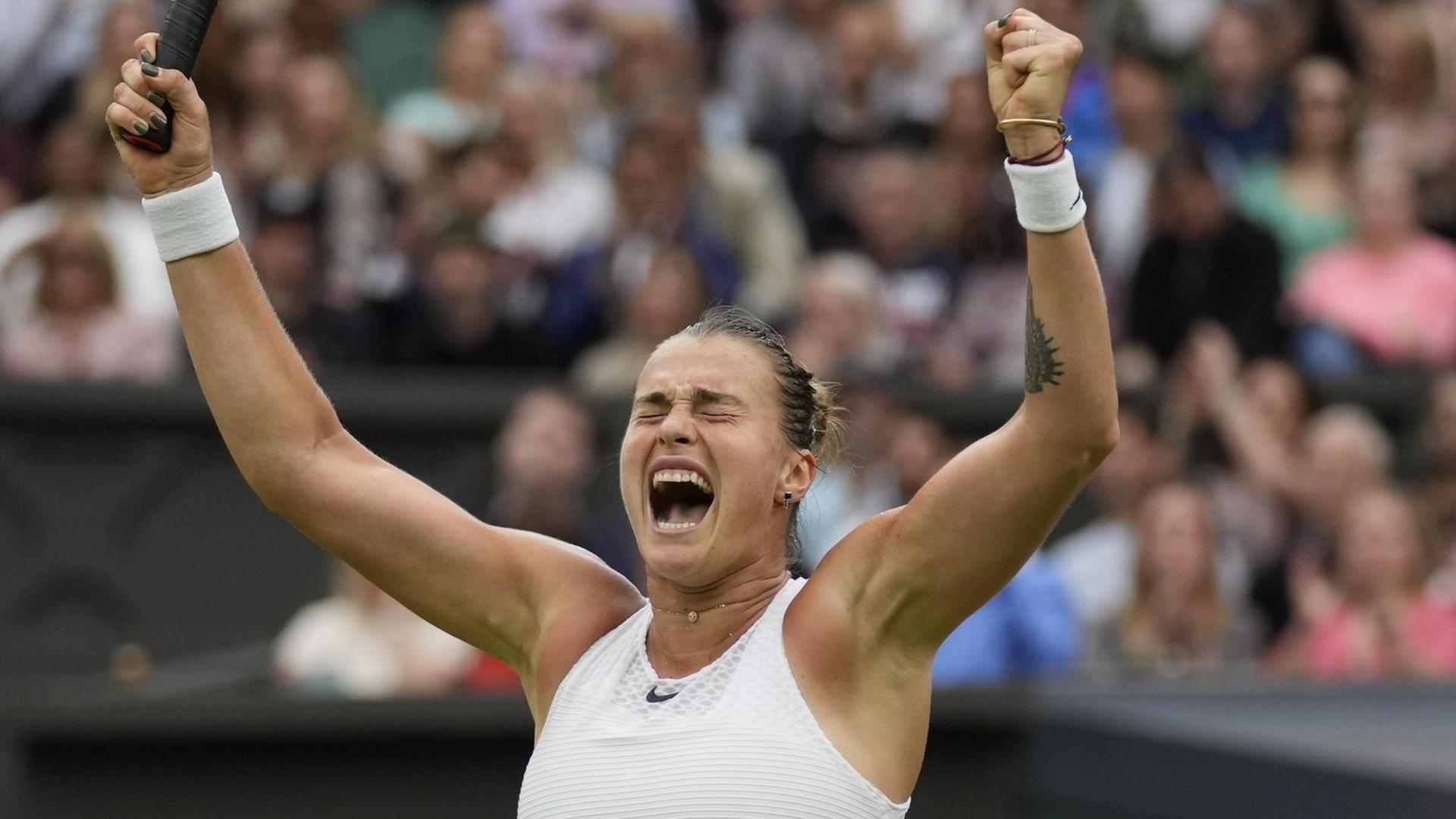 Sabalenka reißt mit zugekniffenen Augen die Arme nach oben und schreit. Im Hintergrund unscharf Zuschauer.