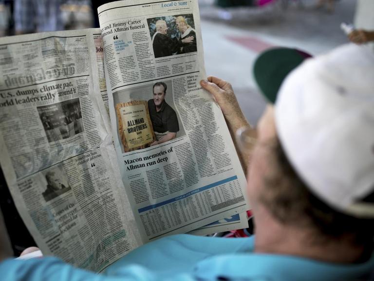 Eine Person mit einer Schirmmütze auf dem Kopf liest eine englischsprachige Zeitung.