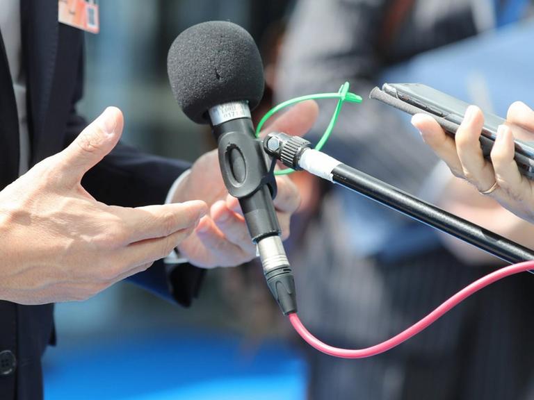 Ein Mikrofon, ein Smartphone und Hände sind in einer Nahaufnahme zu sehen.
