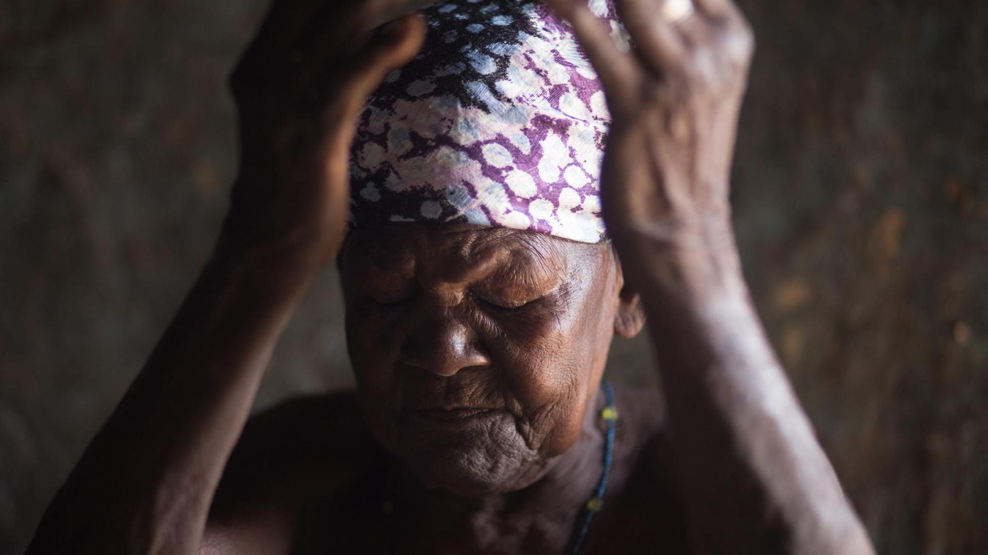 Eine ältere afrikanische Frau, mit einem bunten Kopftuch, hält mit geschlossenen Augen die Hände vor das Gesicht.