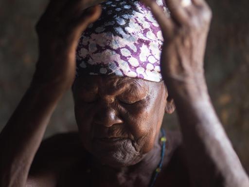 Eine ältere afrikanische Frau, mit einem bunten Kopftuch, hält mit geschlossenen Augen die Hände vor das Gesicht.