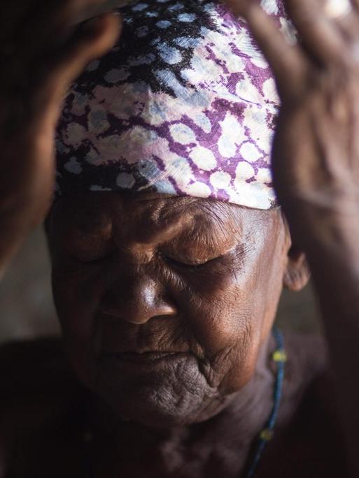 Eine ältere afrikanische Frau, mit einem bunten Kopftuch, hält mit geschlossenen Augen die Hände vor das Gesicht.