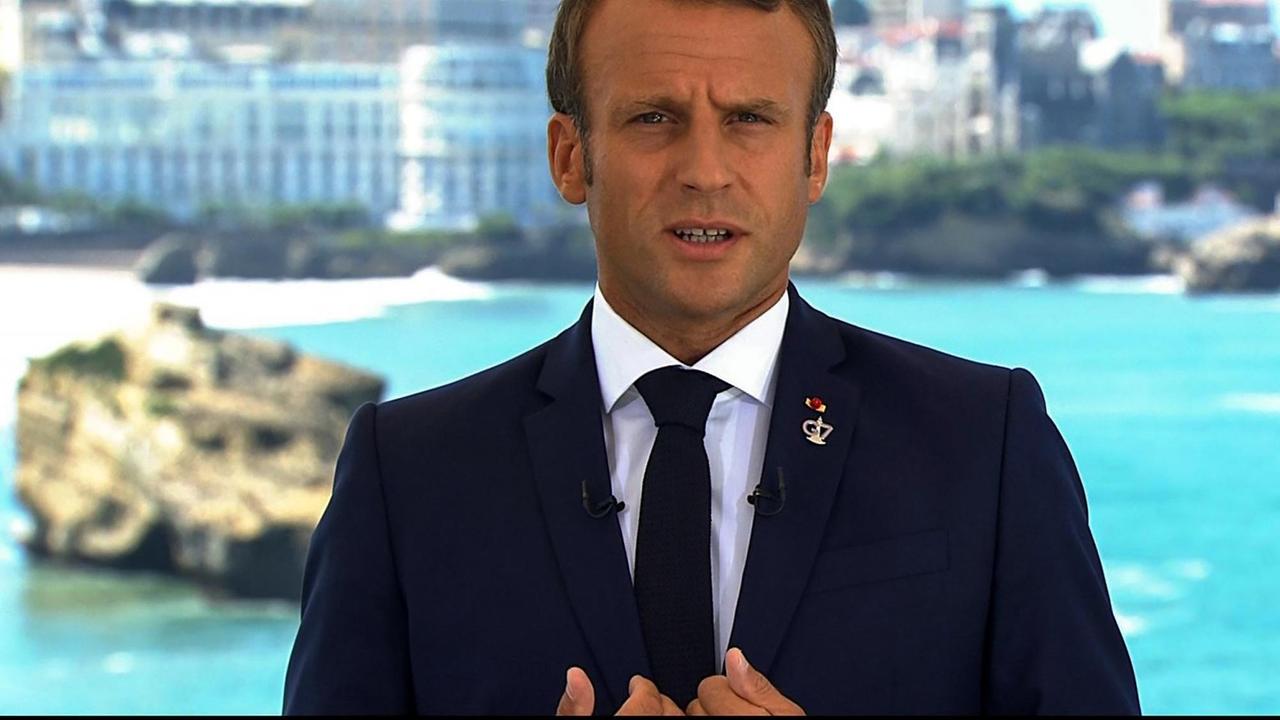 Das Bild zeigt Emmanuel Macron während einer Ansprache auf einem Fernsehbildschirm. Er unterstreicht seine Worte mit gestikulierenden Händen.