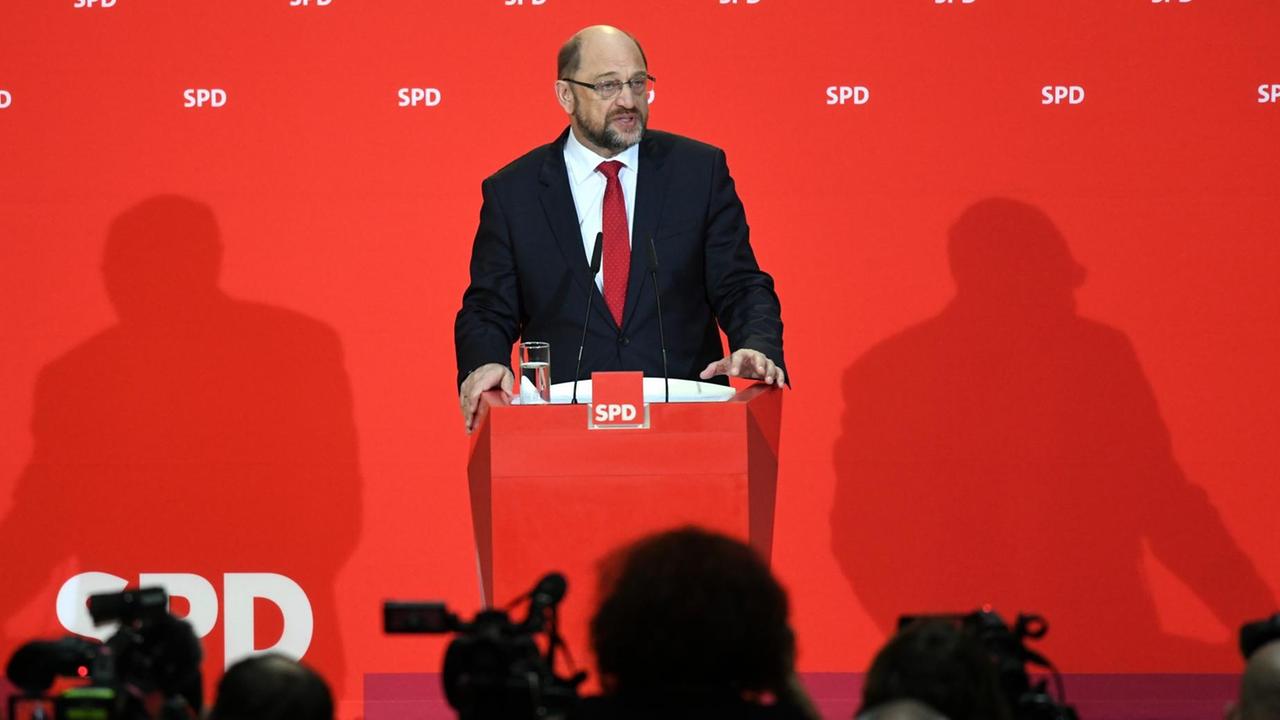 Schulz redet an einem Pult mit Mikrofon vor einer roten Wand mit SPD-Schriftzügen. Im Vordergrund die Köpfe von Journalisten als Silhouetten.