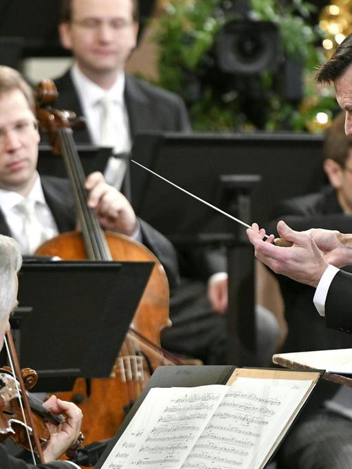 Ein Mann in schwarzem Frack und Taktstock in der rechten Hand ist von seiner linken Seiten aus zu sehen, wie er gebückt vor einem Orchester steht.