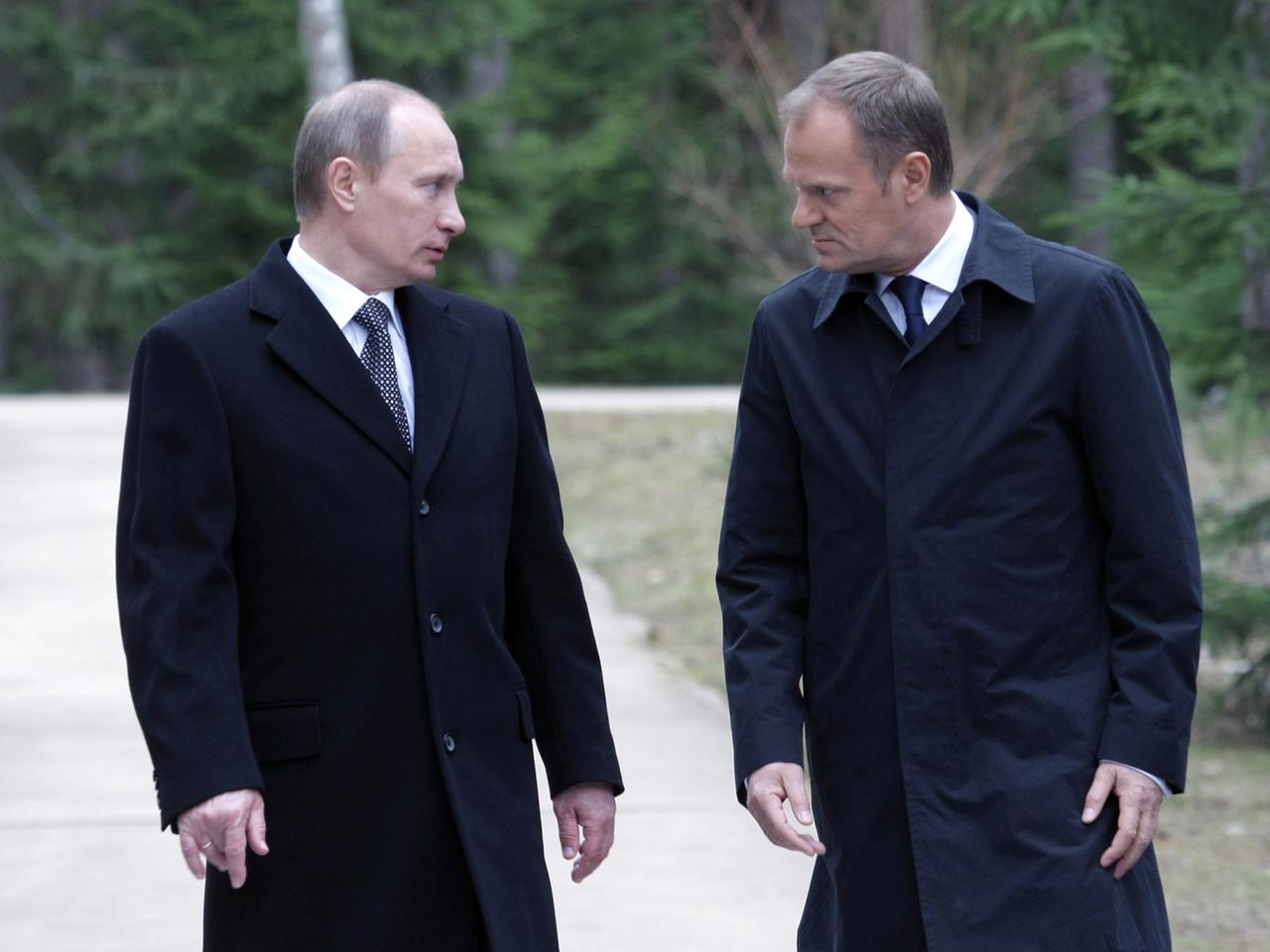 Wladimir Putin und Donald Tusk gehen in schwarzen Mänteln redend nebeneinander her und schauen sich dabei an