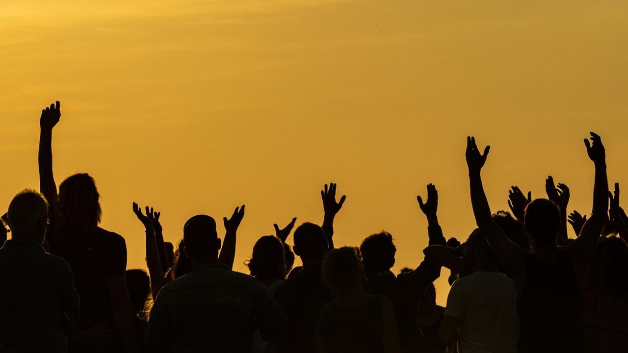 Eine Menschengruppe hebt im Schattenriss die Arme vor einer glutroten Abendstimmung.