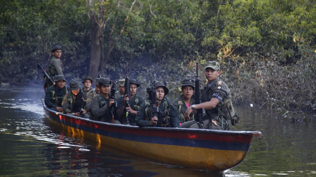 In einem schmalen Boot auf einem Fluss sitzen 12 männliche und weibliche Rebellen in Tarnanzügen und mit Maschinengewehren.