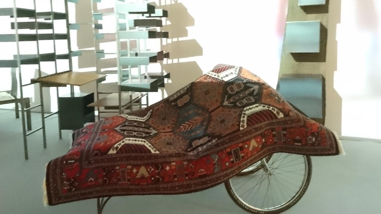 Einblick in die Austellung "Design Gruppe Pentagon" im Museum Angewandte Kunst Köln. Im Vordergrund steht das Objekt "Diwan", das aus einem Untergestell mit zwei großen Speichenrädern, wie bei einer Rikscha, und einem darübergelegten Orientteppich besteht. 