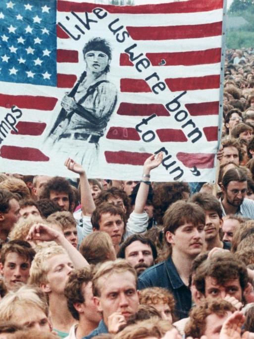 Das Foto vom Publikum beim Bruce-Springsteen-Konzert am 19.07.1988 in Ost-Berlin zeigt begeisterte, junge Männer. Einige Menschen halten ein selbstgemaltes Plakat hoch, darauf eine US-Flagge und die Aufschrift "We Tramps we're born to run" - "Wir Tramps, wir sind geboren, um zu rennen."