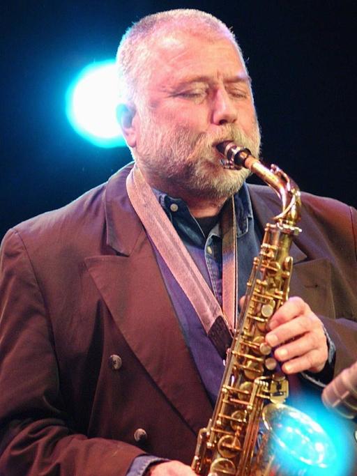 Ein älterer Mann mit kurzgeschorenem weißem Haar und Vollbart spielt mit geschlossenen Augen Altsaxofon. Hinter ihm leuchtet ein blauer Scheinwerfer.