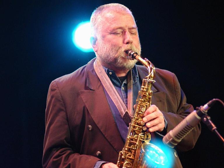 Ein älterer Mann mit kurzgeschorenem weißem Haar und Vollbart spielt mit geschlossenen Augen Altsaxofon. Hinter ihm leuchtet ein blauer Scheinwerfer.