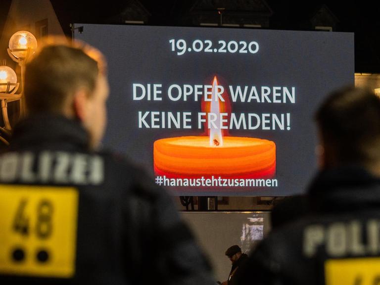 Polizisten stehen auf dem Hanauer Marktplatz, wo auf einer Videoleinwand die Trauerfeier für die Opfer der rassistischen Morde übertragen wird.