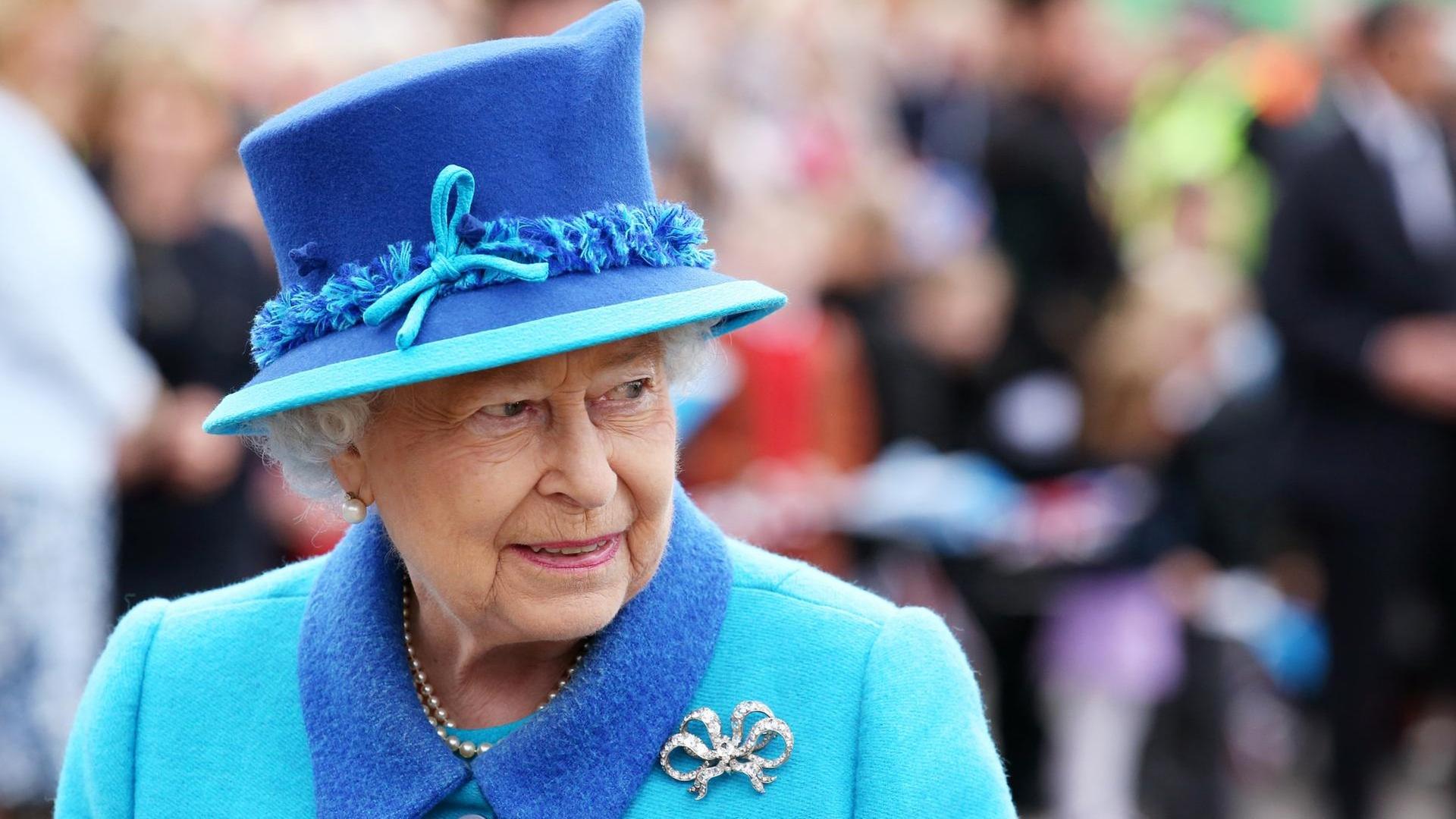 Die Queen steht im hellblauen Kostüm mit dunkelblauem Hut und silberner Brosche im Freien vor einer Menschenmenge, die im Hintergrund unscharf zu erkennen ist.