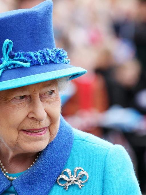 Die Queen steht im hellblauen Kostüm mit dunkelblauem Hut und silberner Brosche im Freien vor einer Menschenmenge, die im Hintergrund unscharf zu erkennen ist.
