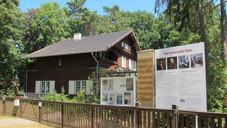 Das dunkelbraune Holzhaus des Komponisten, Pianisten und Dirigenten Xaver Scharwenka in Bad Saarow ist im Hintergrund zu sehen, vorne rechts, hinter dem Holzzaun, eine Infotafel zum Scharwenka Haus.