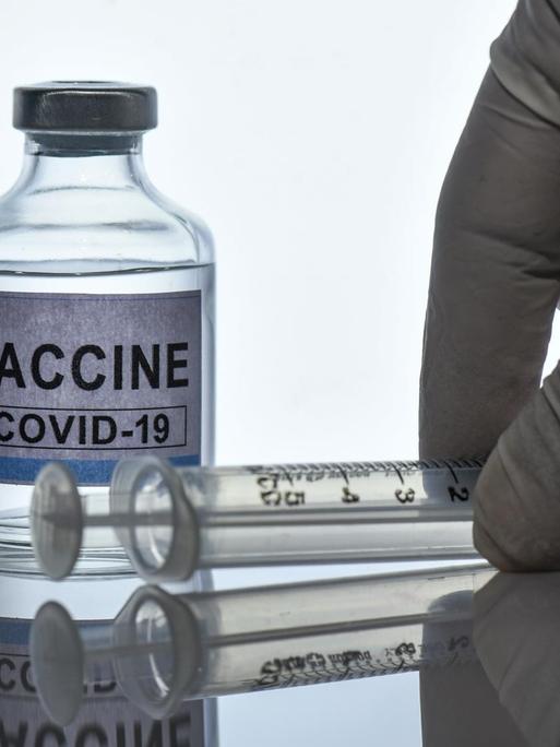 Symbolbild eines Glasfläschchens, auf dem "Vaccine Covid-19" steht, dazu eine behandschuhte Hand und eine Spritze.