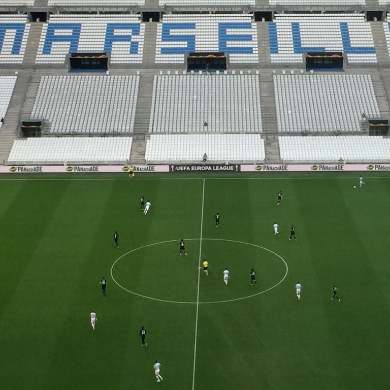 Die Fußballmannschaften von Olympique Marseille und Eintracht Frankfurt spielen, im Hintergrund eine leere Zuschauertribüne mit dem Schriftzug "Marseille".