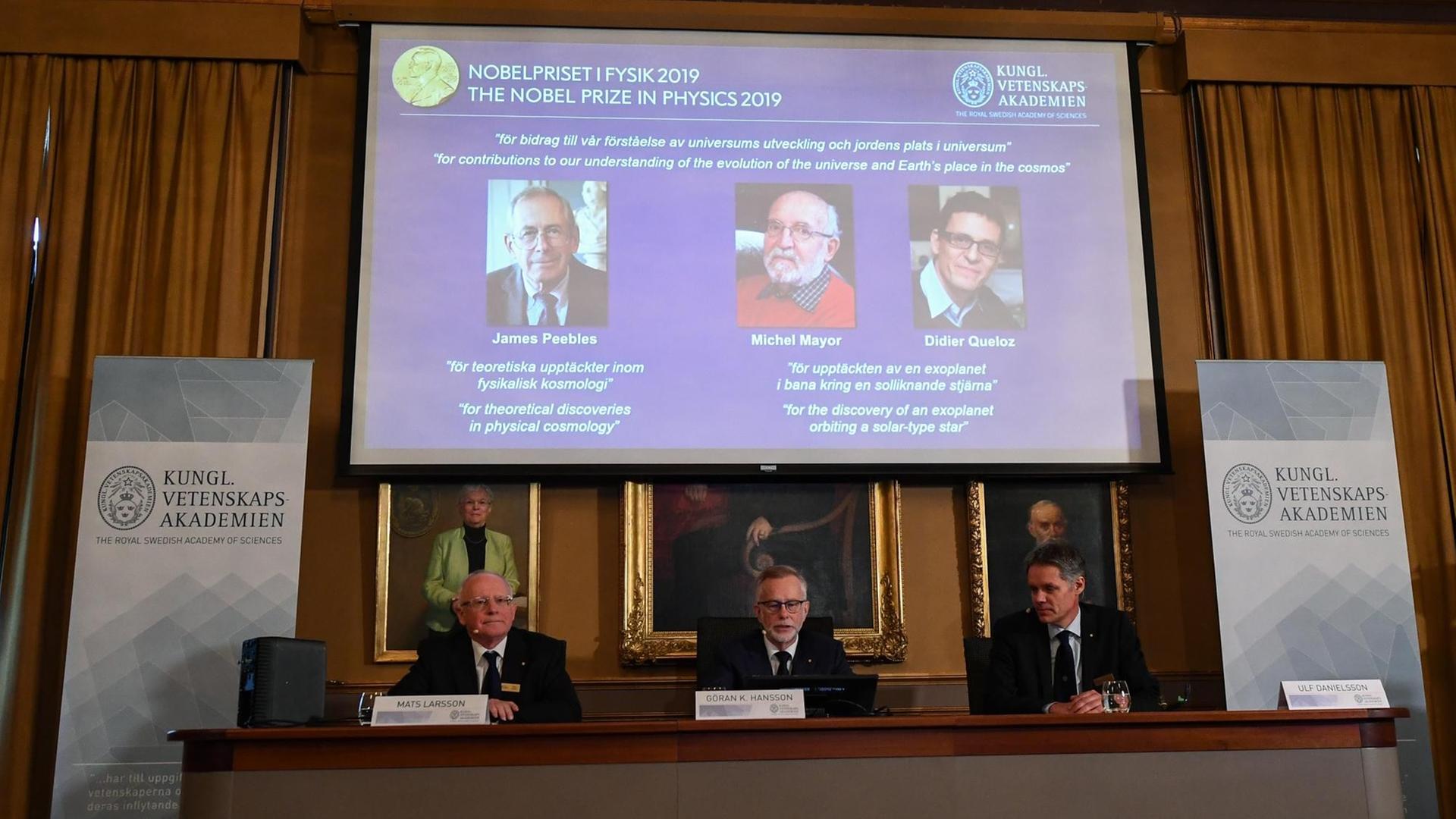 Bekanntgabe der Preisträger in Stockholm. Auf einem Podium sitzen Mitglieder der Schwedischen Akademie der Wissenschaften. Hinter ihnen auf einem Bildschirm sind die Fotos der Preisträger eingeblendet.