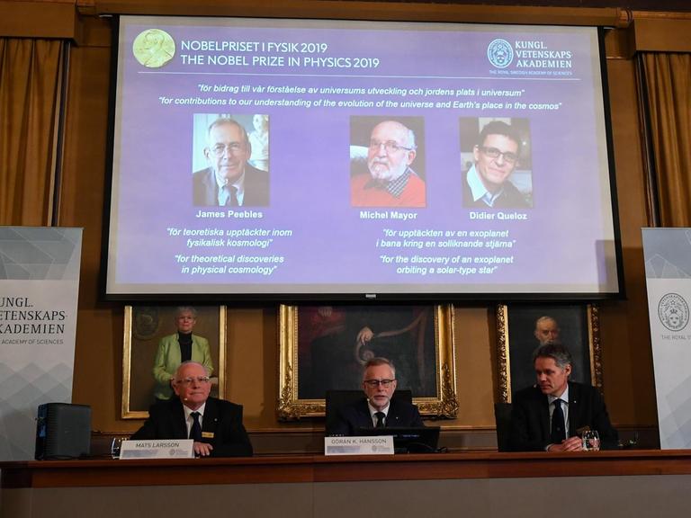 Bekanntgabe der Preisträger in Stockholm. Auf einem Podium sitzen Mitglieder der Schwedischen Akademie der Wissenschaften. Hinter ihnen auf einem Bildschirm sind die Fotos der Preisträger eingeblendet.