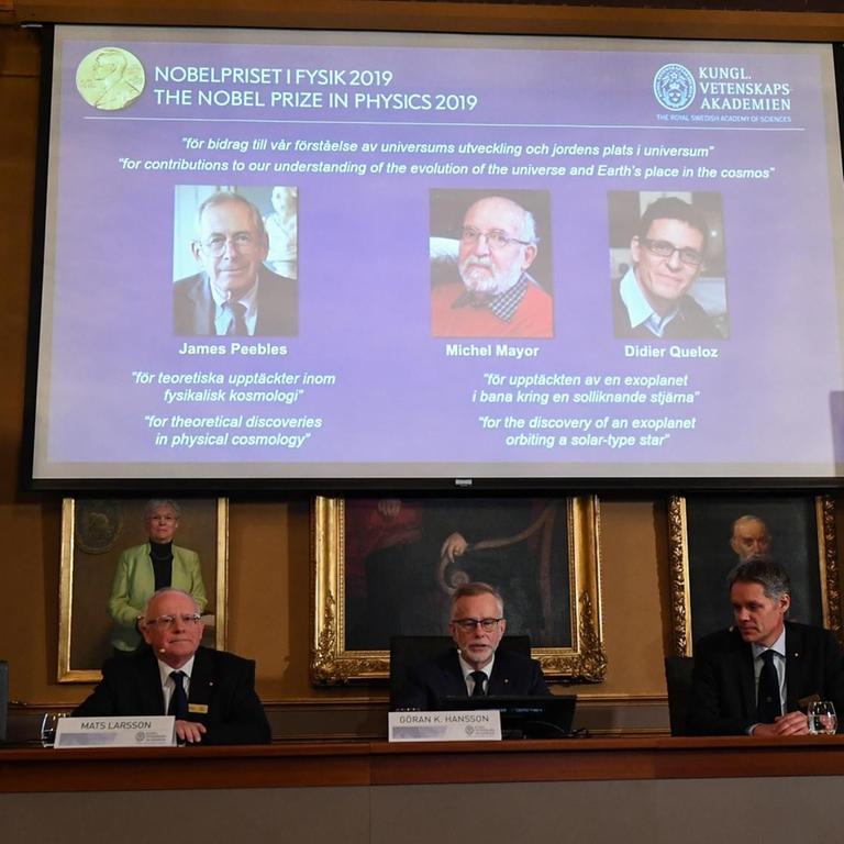 Bekanntgabe der Preisträger in Stockholm. Auf einem Podium sitzen Mitglieder der Schwedischen Akademie der Wissenschaften. Hinter ihnen auf einem Bildschirm sind die Fotos der Preisträger eingeblendet.