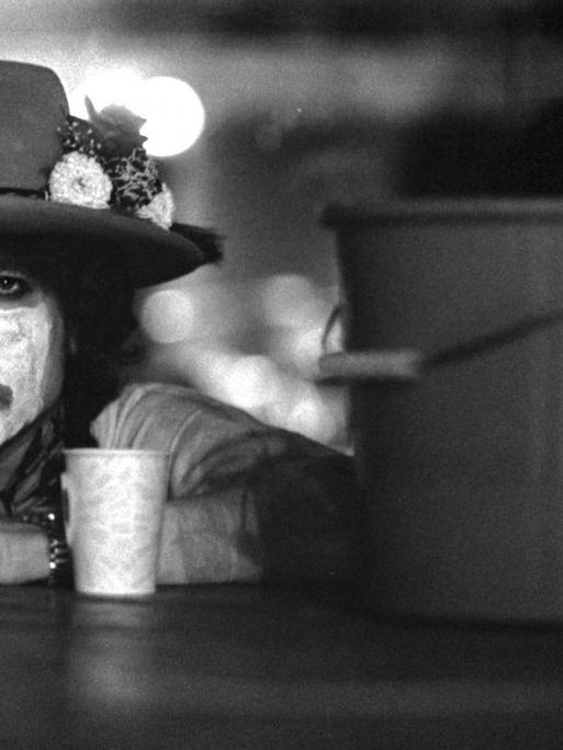 Der Musiker Bob Dylan, die Hände auf einer Tischplatte, mit weiß geschminktem Gesicht und dunklem Hut sowie mit einem Kaffeebecher vor und einem Putzeimer neben sich.