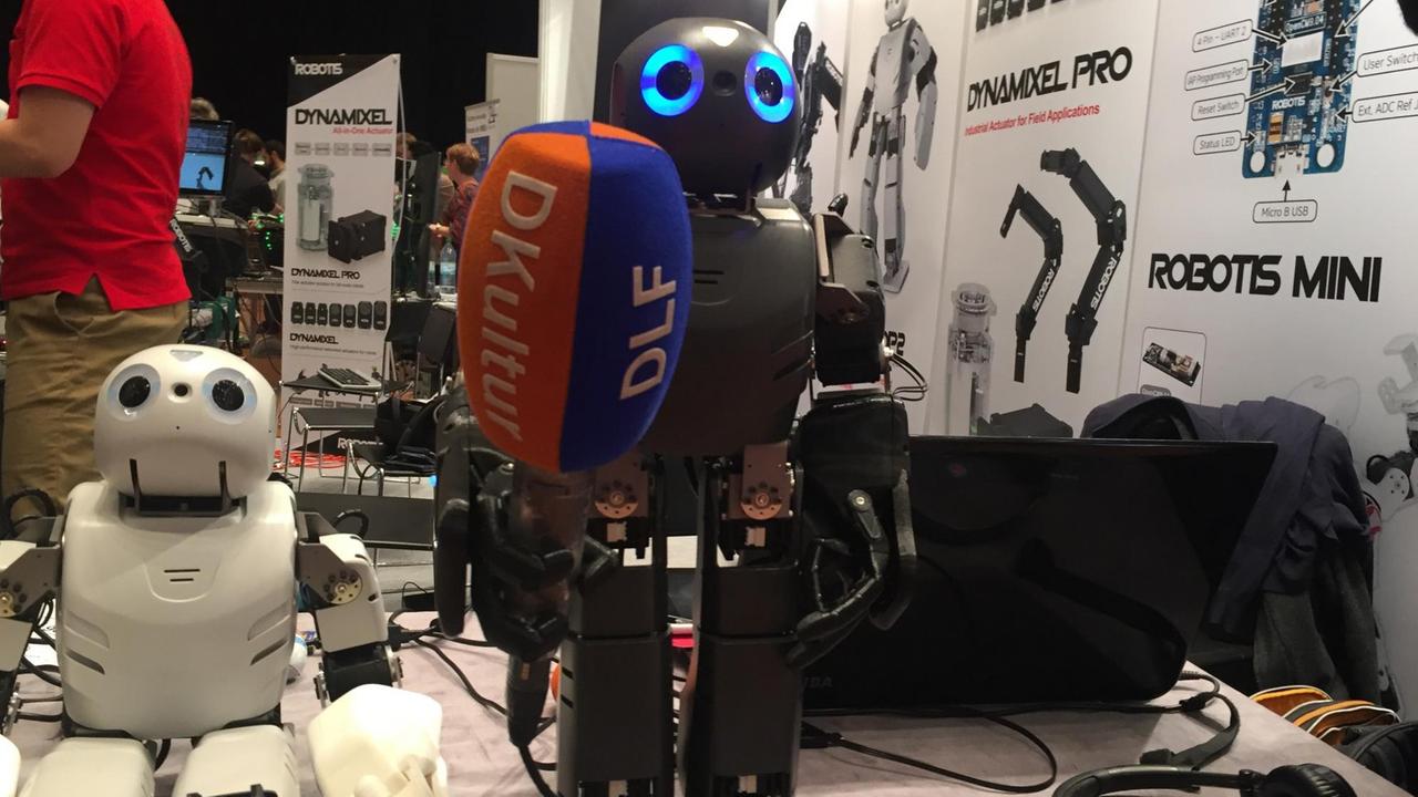Zwei Roboter, fotografiert auf der Roboter Messe Hamburg. Einer hält ein Mikrofon in der Hand. 