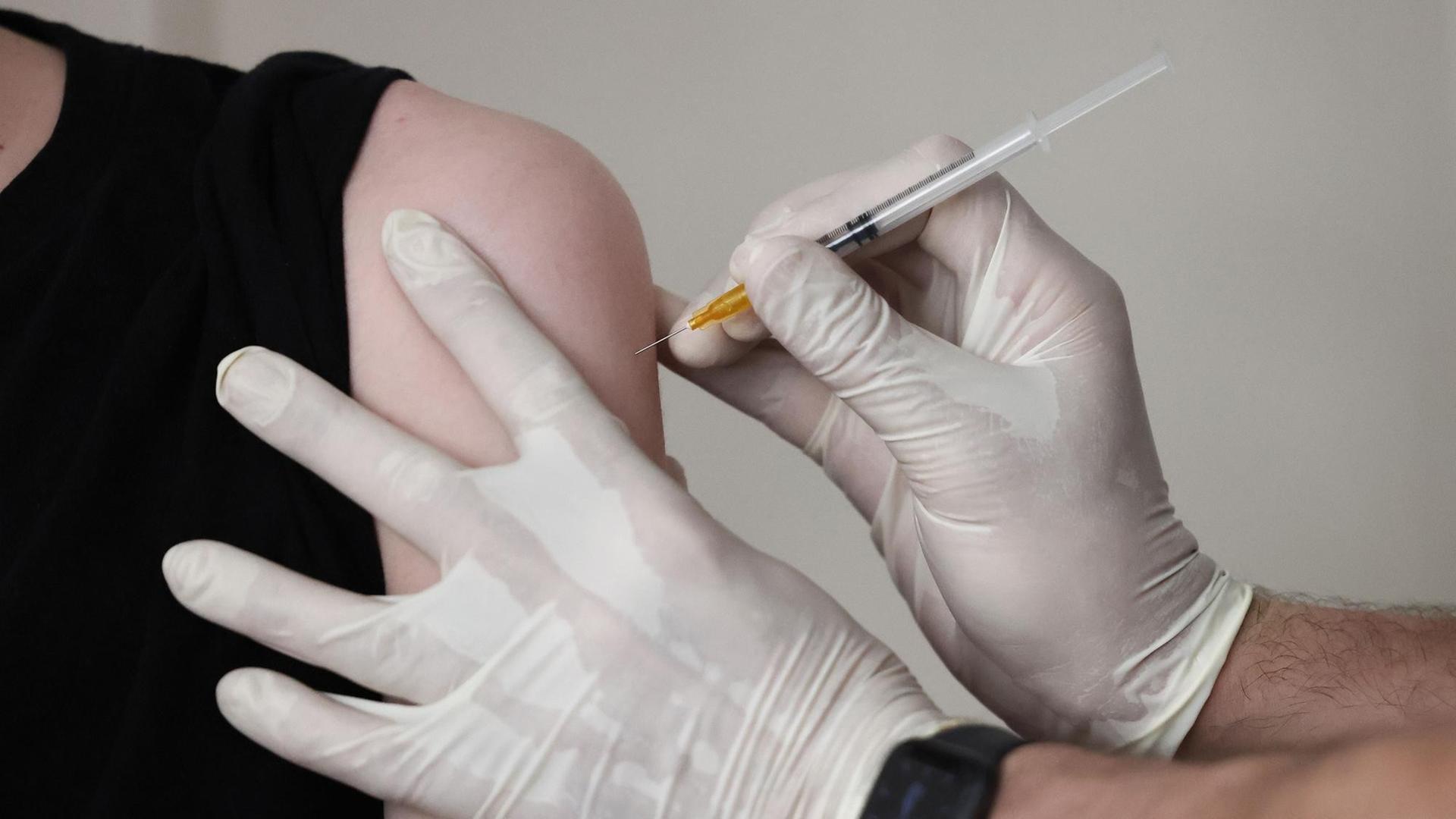 Zu sehen sind die Hände eines Arztes mit medizinischen Handschuhen, der ein Vakzin in einer Spritze in den Oberarm eines Patienten verabreicht. Zu sehen sind die Hände eines Arztes mit medizinischen Handschuhen, der ein Vakzin in einer Spritze in den Oberarm eines Patienten verabreicht.