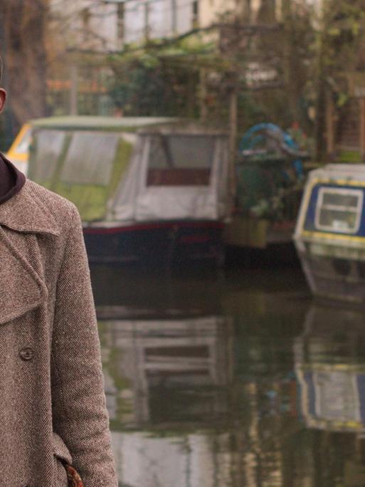 Porträt von JJ Bola, der im Mantel vor einem Kanal mit Boten im Hintergrund steht.