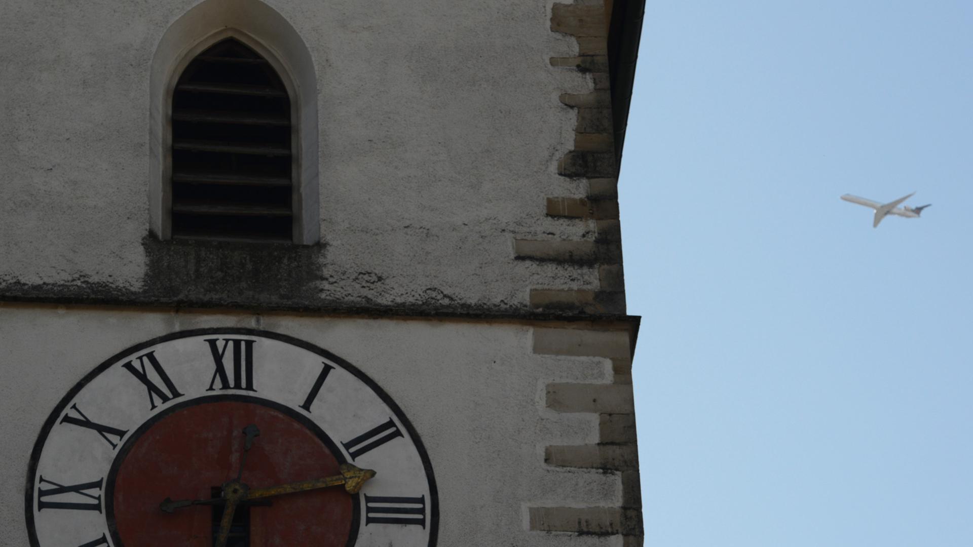 Die Uhr eines Kirchturms in Großaufnahme, daneben weit hinten ein Flugzeug im Landeanflug vor blauem Himmel.