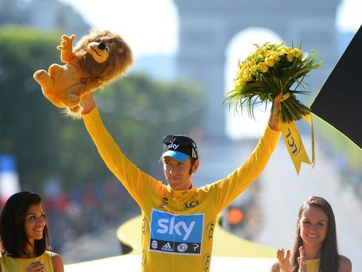 Bradley Wiggins trägt ein gelbes Trikot und reckt in Siegerpose beide Arme in die Höhe. In der rechten Hand hat er einen Plüsch-Löwen, in der linken einen Strauß mit gelben Blumen.