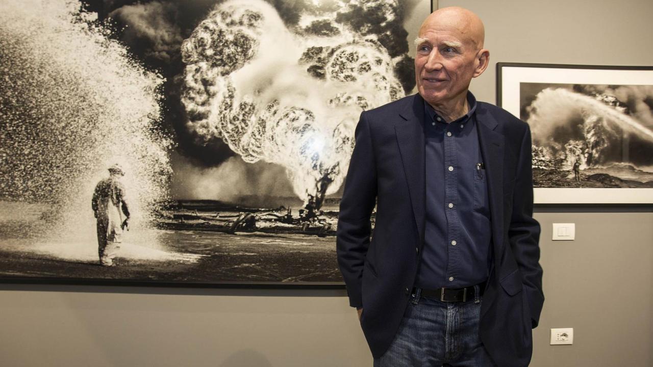 Der Fotograf Sebastião Salgado steht bei der Eröffnung seiner "Genesis"-Ausstellung 2017 in Mailand vor einer seiner Fotografien. Zu sehen ist eine Schwarzweißaufnahme eines brennenden Ölfeldes in Kuwait mit einem Arbeiter. Der Fotograf Sebastião Salgado steht bei der Eröffnung seiner "Genesis"-Ausstellung 2017 in Mailand vor einer seiner Fotografien. Zu sehen ist eine Schwarzweißaufnahme eines brennenden Ölfeldes in Kuwait mit einem Arbeiter.